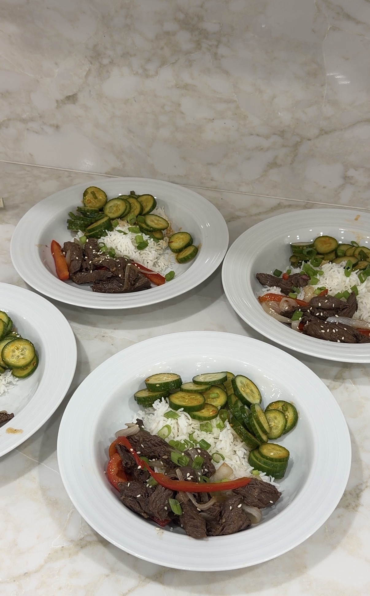 a bowl of sliced skirt steak with jasmine rice, peppers, onions, scallions, sesame seeds, and chili crisp cucumber salad